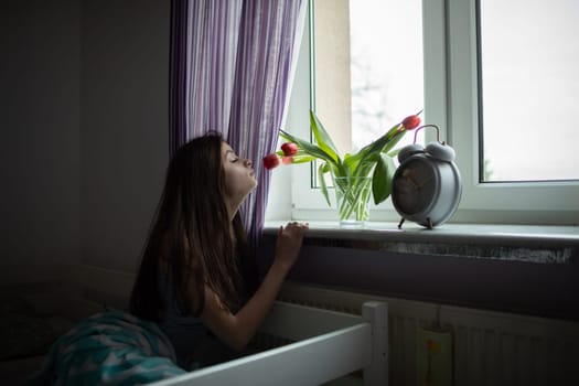 A girl sniffs the scent of flowers standing on her windowsill. Bright bedroom with decorations.