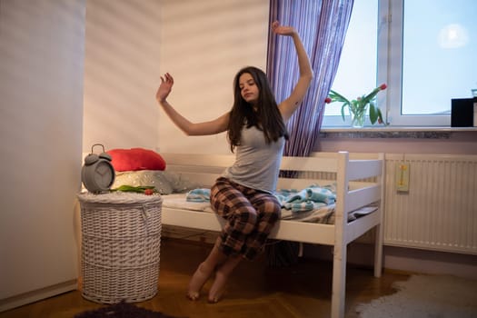 A teenage girl with long hair stretches sitting on the edge of the bed. Bright bedroom with decorations.