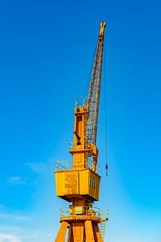 Old and obsolete yellow crane on the harbor pier lit by the afternoon sun with blue sky in the background.