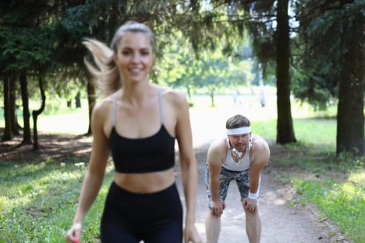 Beautiful athlete woman and tired man behind in park on jog. Sport health and fatigue concept