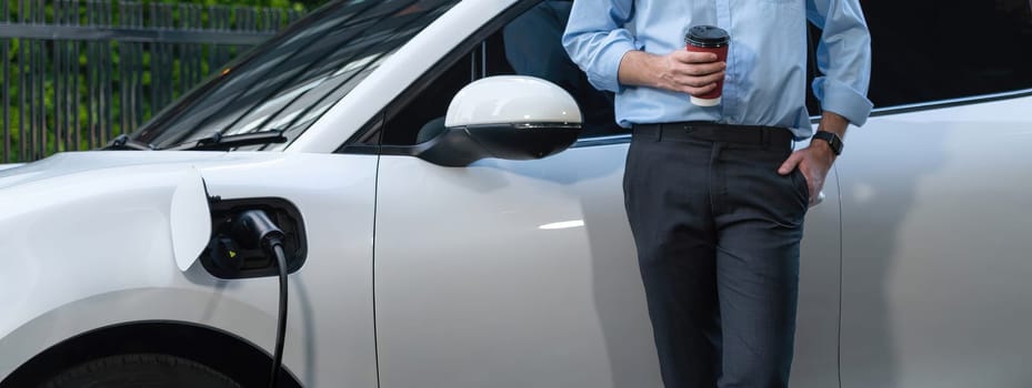 Progressive eco-friendly concept of parking EV car at public electric-powered charging station in city with blur background of businessman leaning on recharging-electric vehicle with coffee.