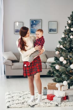 Beautiful young mother holds her little son by hands at home near New year christmas tree.
