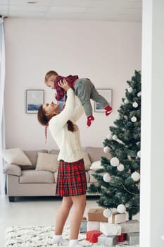 Beautiful young mother holds her little son by hands at home near New year christmas tree.