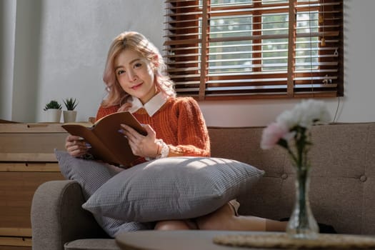 Attractive young Asian woman sits in the minimal and comfortable living room enjoying reading a book...