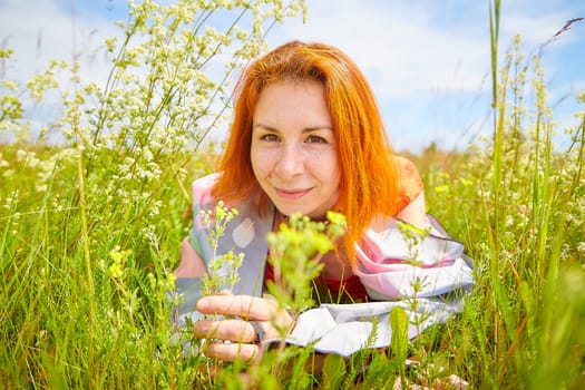 Outdoors portrait of red haired woman in grass and lower on field and meadow. Young woman enjoying her freedom in flowering nature