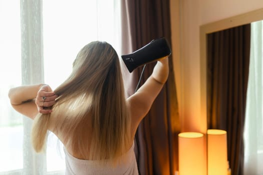 Happy woman drying her hair in bathroom. Hair care. Beautiful smiling woman drying healthy hair using hair dryer. Girl in bath towel is using a dryer and smiling while looking into the mirror
