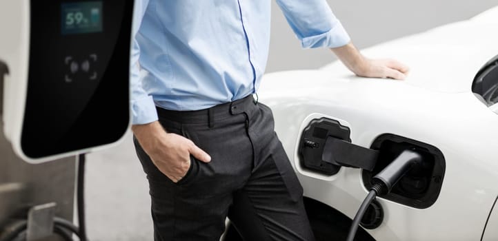 Closeup progressive suit-clad businessman with his electric vehicle recharge his car on public charging station in modern city with power cable plug and renewable energy-powered electric vehicle.