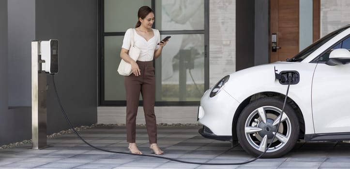 Progressive asian woman holding smartphone with electric car at home charging station. Concept of the use of electric vehicles in a progressive lifestyle contributes to a clean and healthy environment