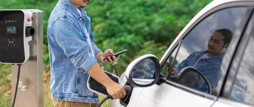 Progressive man with his electric car, EV car recharging energy from charging station on green field with wind turbine as concept of future sustainable energy. Electric vehicle with energy generator.