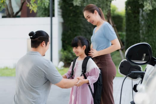 Progressive young parents and daughter with electric vehicle and home charging station. Green and clean energy from electric vehicles for healthy environment. Eco power from renewable source at home.