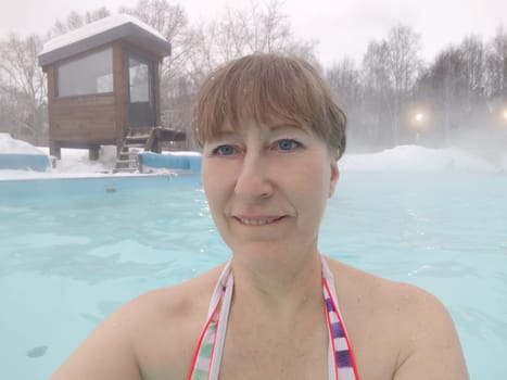 Adult mature woman takes selfie in a pool with warm hot termal mineral water in winter and white snow around. Wellness center and the concept of health care. Travel, recreation, medicine and rest