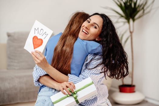 Happy mother's day. Child daughter congratulates mom and gives her postcard. Mum and girl smiling and hugging. Family holiday and togetherness. Mom is happy to receive an unexpected gift from her child.