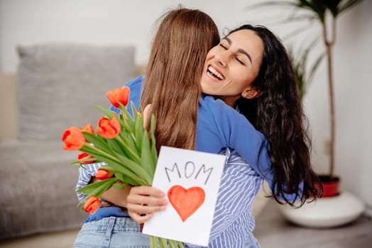Child daughter congratulates mom and gives her flowers tulips and postcard. Mum and girl smiling and hugging. Family holiday and togetherness. Mom hugs her daughter in her arms, holds her gifts and closed her eyes in pleasure.