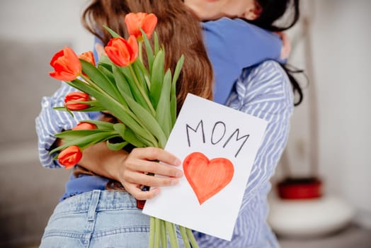 Grateful mom hugging daughter girl, holding flowers bouquet, receiving hand drawn greeting card with loving heart from girl, smiling at camera. Mothers day, 8 march, concept. Head shot portrait. Close-up of hands holding a bouquet of tulips and a handmade card.