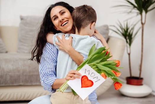 Little boy greeting his mom on Mother's Day at home. The son gave his mother a bouquet of flowers and a handmade card.Mom hugs her child and smiles broadly.