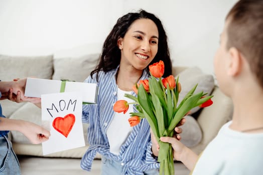 Arab Mom receives gifts from her children. The son holds out a bouquet of tulips and the daughter a handmade card and a gift. Everyone is in a bright room in a cozy homely atmosphere.