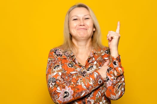 Caucasian blonde woman in a patterned dress raising her index finger smiling, she had a great innovative idea that solves the problem. Isolated on yellow background