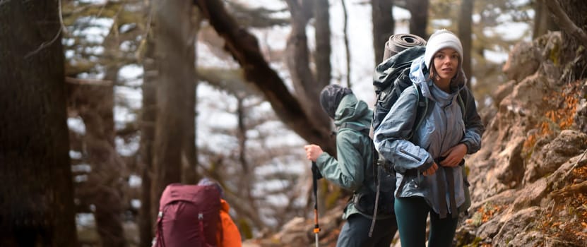 Young stylish travelers discover and enjoy the nature, walk the trail among the mountains and forest. Three girls spend their time actively and go hiking with good tourist equipment.
