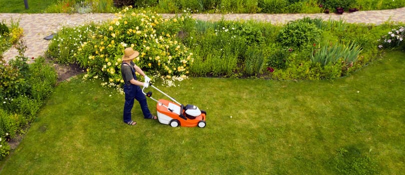 A young man is mowing a lawn with a lawn mower in his beautiful green floral summer garden. A professional gardener with a lawnmower cares for the grass, view from above