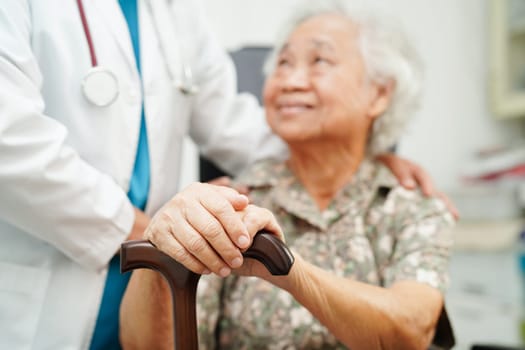 Doctor help Asian elderly disability woman patient holding walking stick in wrinkled hand at hospital.