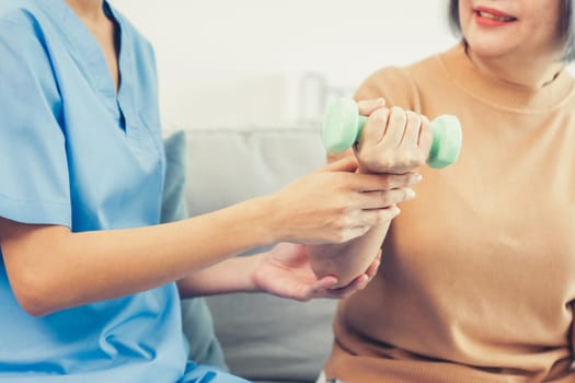 Caregiver helping contented senior woman exercise with dumbbell at home. Rehab for elder, senior medical care.