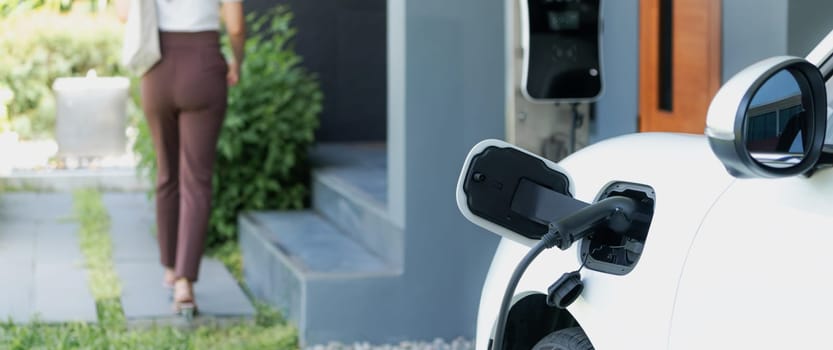 Focus EV charger plugged into EV car at home charging station with blurred background of progressive woman walking in background. Elective vehicle powered by clean energy for eco-friendly concept.