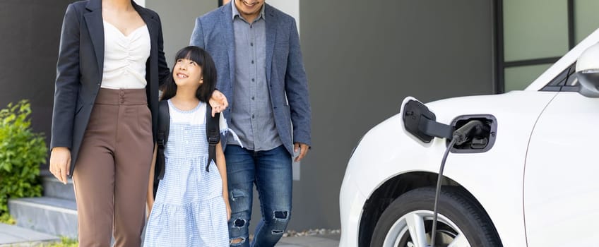 Progressive young parents and daughter with electric vehicle and home charging station. Green and clean energy from electric vehicles for healthy environment. Eco power from renewable source at home.