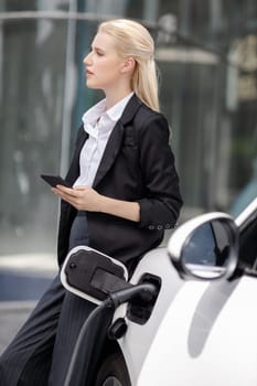 Businesswoman wearing black suit using smartphone, leaning on electric car recharge battery at charging station in city residential building with condos and apartment. Progressive lifestyle concept.