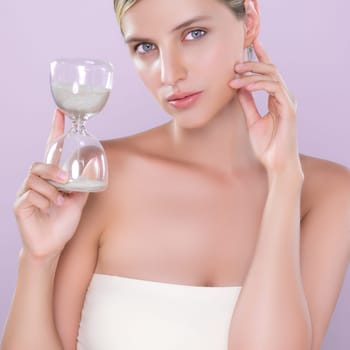 Alluring beauty model using hourglass in beauty concept of anti-aging skincare treatment for woman. Beautiful caucasian women portrait with perfect smooth clean skin in pink isolated background.