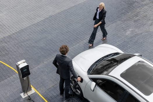 Aerial view progressive lifestyle of businesspeople with electric car connected to charging station at city center public parking car. Eco friendly rechargeable car powered by alternative clean energy