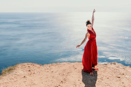 Side view a Young beautiful sensual woman in a red long dress posing on a rock high above the sea during sunrise. Girl on the nature on blue sky background. Fashion photo.