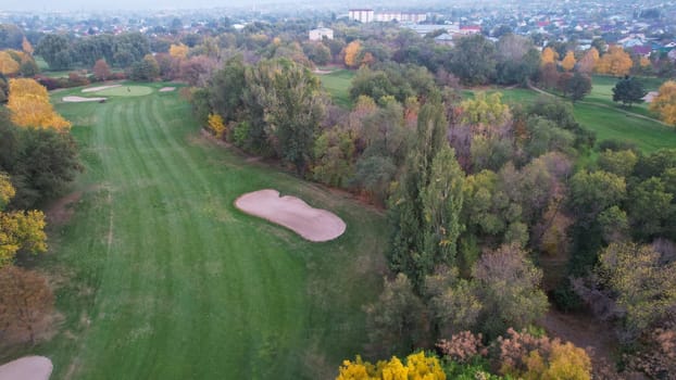 People play golf on a green field in autumn. Yellow-red leaves on some trees. Places for holes. There is a golf car. Houses and mountains are visible in the distance in a haze. Sunset. Wedding