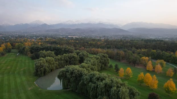 People play golf on a green field in autumn. Yellow-red leaves on some trees. Places for holes. There is a golf car. Houses and mountains are visible in the distance in a haze. Sunset. Wedding