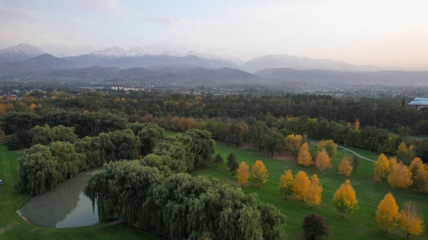 People play golf on a green field in autumn. Yellow-red leaves on some trees. Places for holes. There is a golf car. Houses and mountains are visible in the distance in a haze. Sunset. Wedding