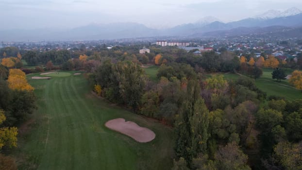 People play golf on a green field in autumn. Yellow-red leaves on some trees. Places for holes. There is a golf car. Houses and mountains are visible in the distance in a haze. Sunset. Wedding