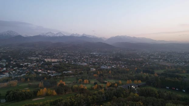 People play golf on a green field in autumn. Yellow-red leaves on some trees. Places for holes. There is a golf car. Houses and mountains are visible in the distance in a haze. Sunset. Wedding