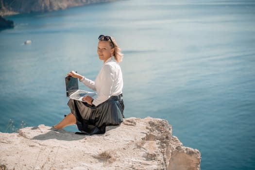 Business woman on nature in white shirt and black skirt. She works with an iPad in the open air with a beautiful view of the sea. The concept of remote work