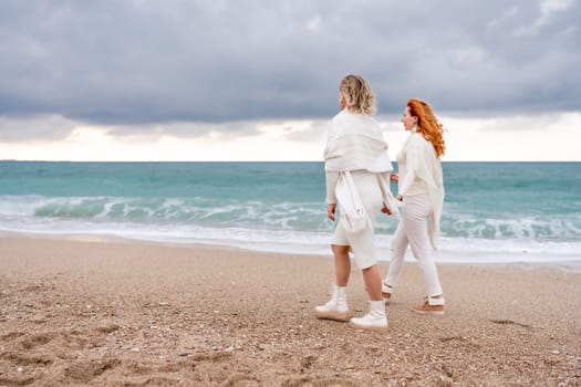 Women sea walk friendship spring. Two girlfriends, redhead and blonde, middle-aged walk along the sandy beach of the sea, dressed in white clothes. Against the backdrop of a cloudy sky and the winter sea. Weekend concept