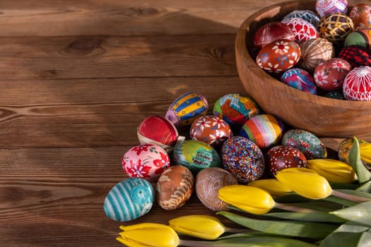Intricately arranged Easter eggs on a wooden tabletop and in a wooden bowl. The Easter composition is completed with swishy yellow tulips.