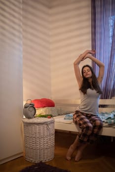 A woman in pajamas sits on the bed and raises her arms up. The light of a lamp on the wall.