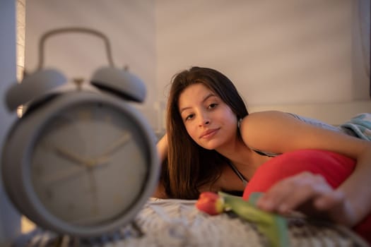 The brunette reaches for a flower on the nightstand. The light of a lamp on the wall. A large alarm clock stands on the bedside table.