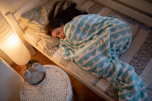 The tired girl sleeps wrapped in a blanket. The light of a lamp on the wall.