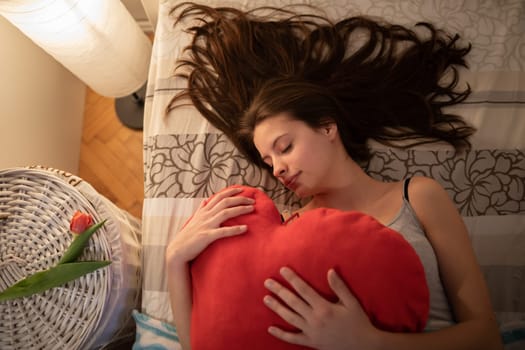 A teenage girl hugs a red heart-shaped pillow. The light of a lamp on the wall.