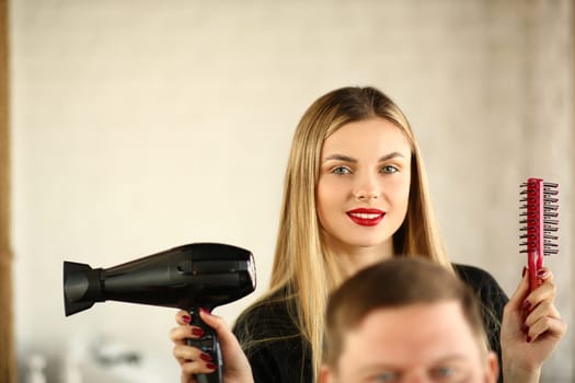Woman hairdresser is holding hair dryer and comb man is sitting in chair. Stylish styling of men hair concept