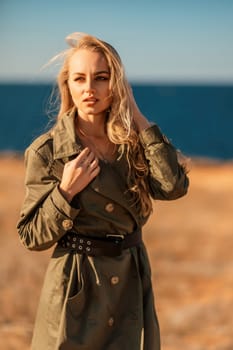 Portrait blonde sea cape. Calm young blonde in a khaki raincoat stands on the seashore.