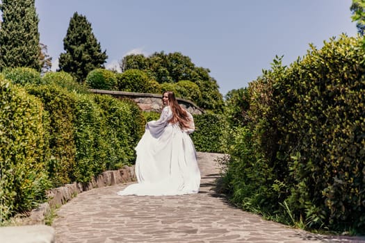 Brunette runs white dress park. A beautiful woman with long brown hair and a long white dress runs along the path along the beautiful bushes in the park, rear view.