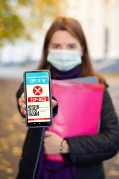 Young girl in medical mask standing near university building showing to the camera red expired electronic certificate