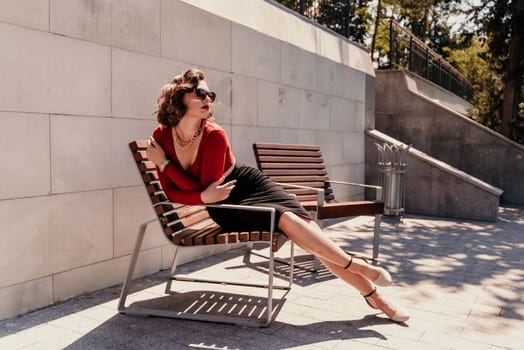 Portrait of a woman on the street. An attractive woman in glasses, a red blouse and a black skirt is sitting on a bench outside