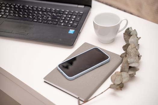 White desktop with laptop, coffee mug, phone and notebook for remote work or study. Office computer workplace in home interior.