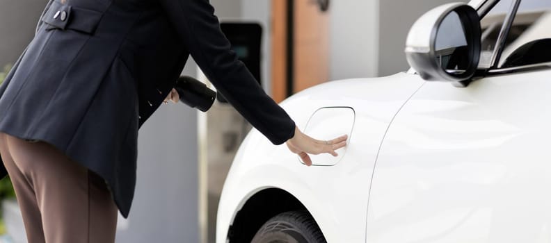 Closeup progressive woman install cable plug to her electric car with home charging station. Concept of the use of electric vehicles in a progressive lifestyle contributes to clean environment.